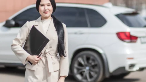 Uśmiechnięta kobieta w eleganckim płaszczu trzymająca teczkę, stojąca przed białym samochodem